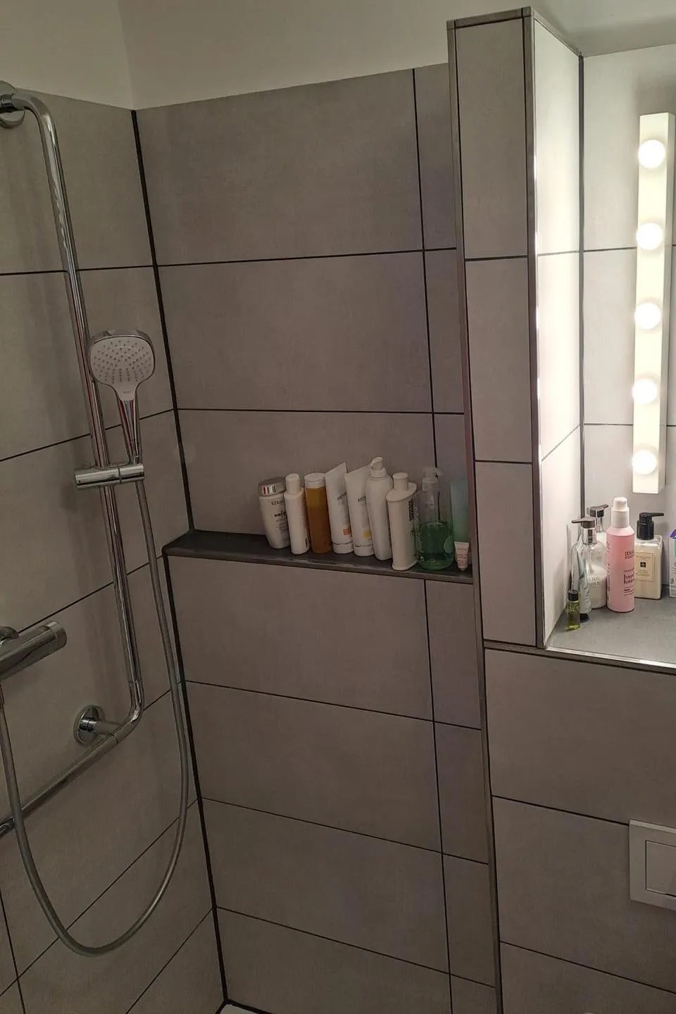 Fertiges Bad unter der Dachschräge mit großformatigen grauen Fliesen, gefliester Ablagenische in der Dusche, Thermostat-Brausegarnitur und beleuchtetem Spiegel über einer gefliesten Vorwand.